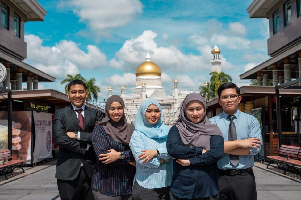 A team of confident professionals from Brunei Energy Services & Trading Company standing proudly in front of a beautiful mosque with a golden dome under a clear blue sky.