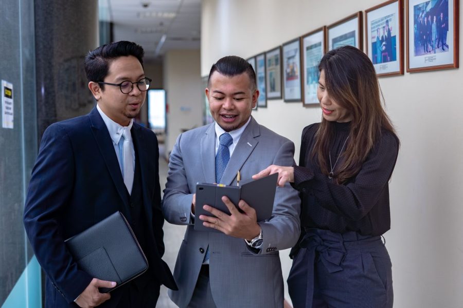 Three professionals in business attire engaged in a focused discussion over a digital tablet in an office hallway at Brunei Energy Services & Trading Company.