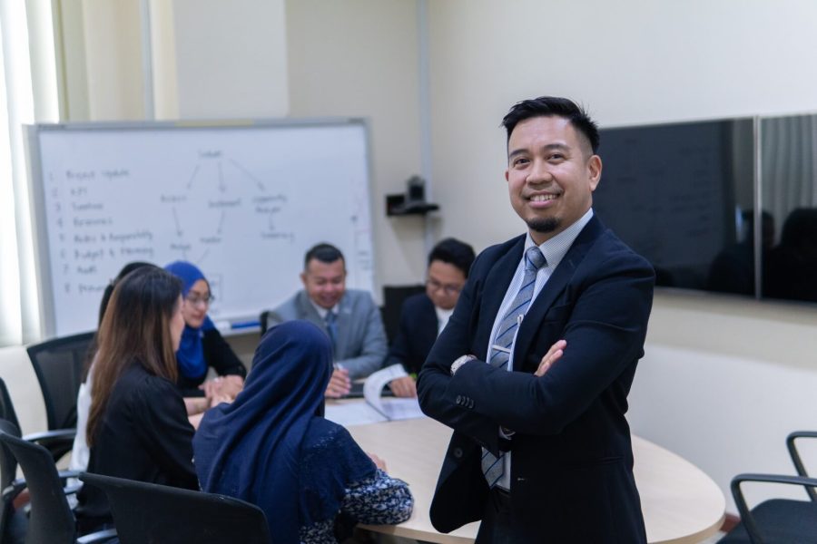 Confident team leader from Brunei Energy Services & Trading Company smiling in a business meeting with colleagues discussing in the background.